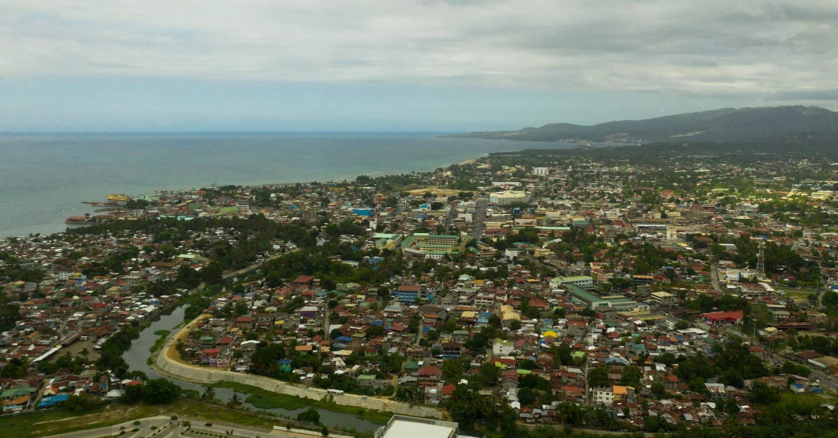 Aerial view of Iligan City with densely packed buildings, a winding river, and a mountainous backdrop.