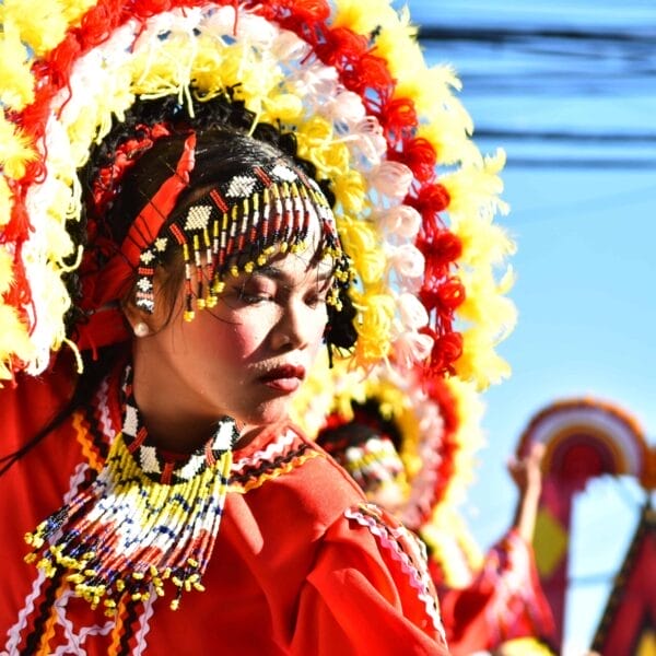 Dancer in red costume, colorful headdress, eyes closed, celebrating Diyandi Festival against a bright blue sky.