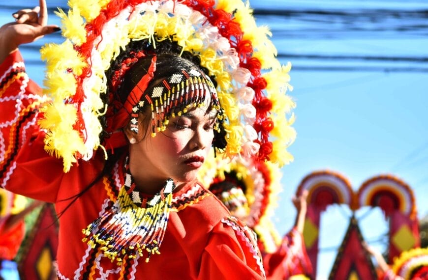 Dancer in red costume, colorful headdress, eyes closed, celebrating Diyandi Festival against a bright blue sky.