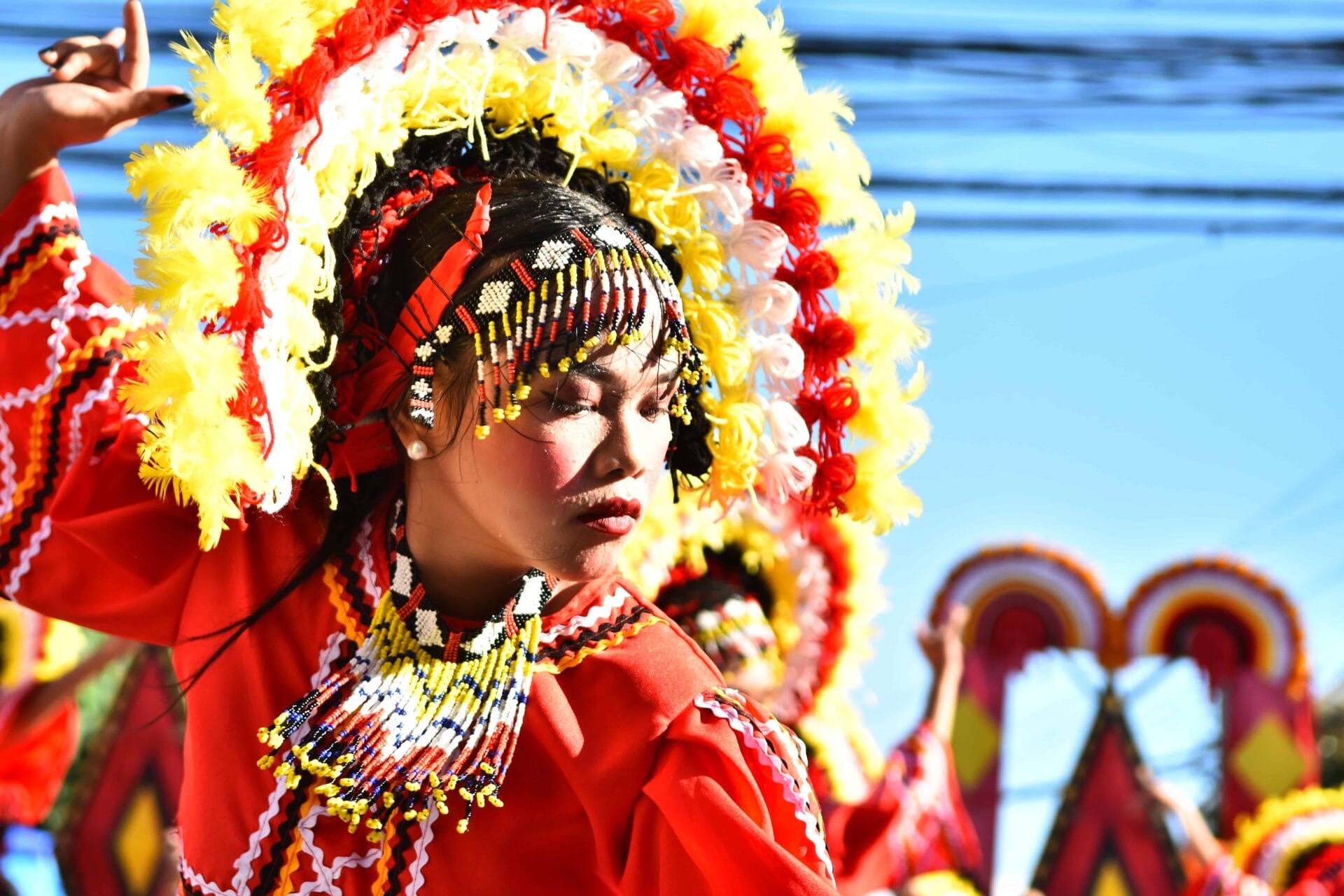 Dancer in red costume, colorful headdress, eyes closed, celebrating Diyandi Festival against a bright blue sky.