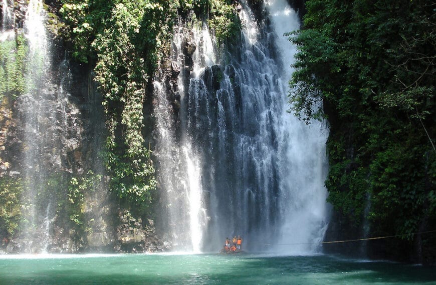 Spectacular Tinago Falls cascading into a turquoise pool, lush greens enveloping; people in orange vests below.