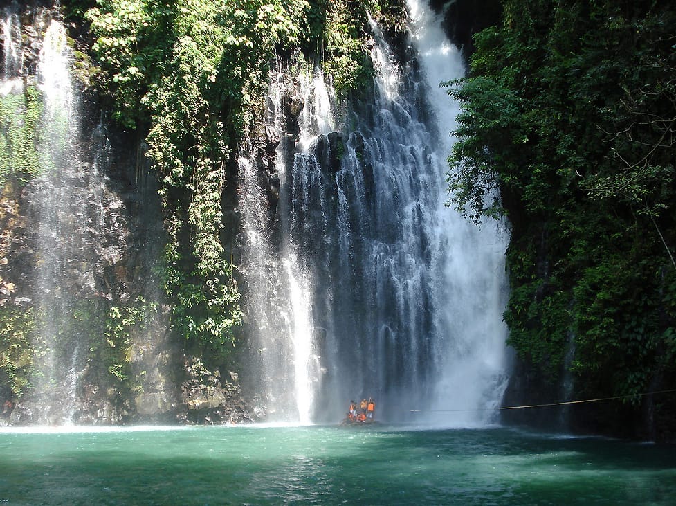 Spectacular Tinago Falls cascading into a turquoise pool, lush greens enveloping; people in orange vests below.