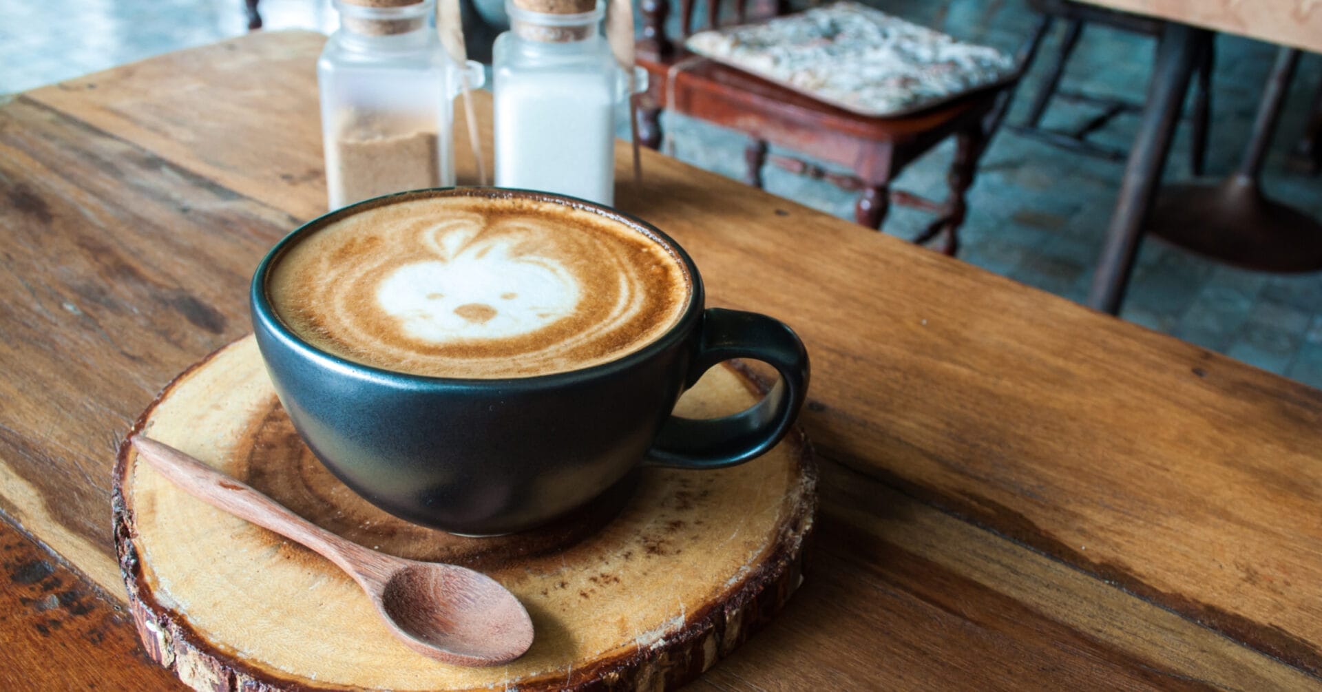 Latte art of a rabbit in a dark cup at Iligan Coffee, with sugar jars and a chair on a wooden coaster.